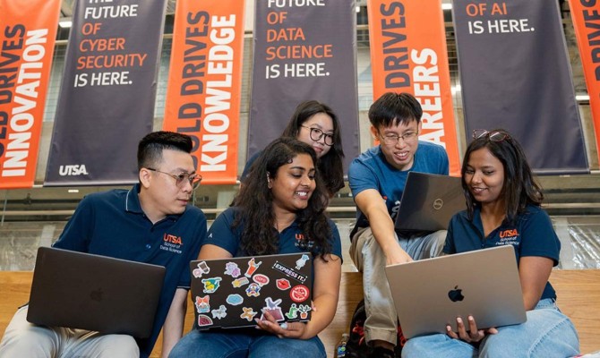 A group of students in blue shirts sit on a staircase looking at their laptops.