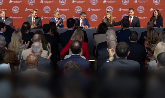 A panel of UT Austin leadership, Board Chairman and Michael and Susan Dell sit at a long table on a stage with a orange backdrop in repeating logos. 