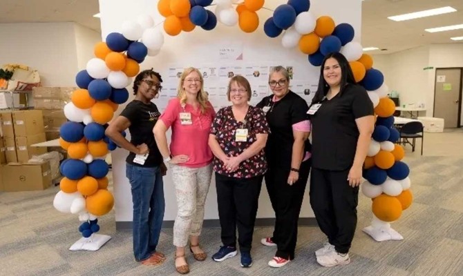 Five nurse women wearing name badges stand together smiling beneath a balloon arch
