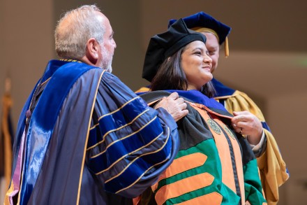Image of a female student graduating and receiving her doctoral hood from two male, robed faculty members