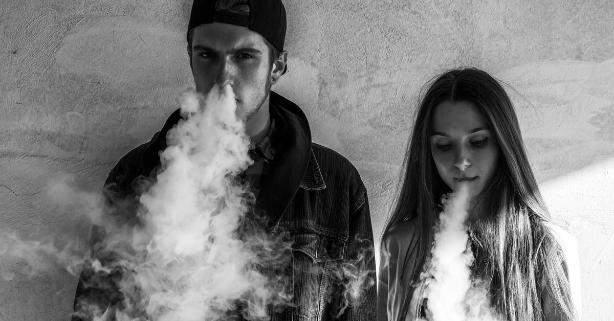 Two young adults, a man and woman, exhaling vaping trail smoke